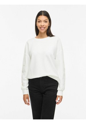 Smilende kvinne med langt mørkt hår iført en hvit langermet topp med blomsterutskjæringer på ermene og svarte, høytlivede bukser.