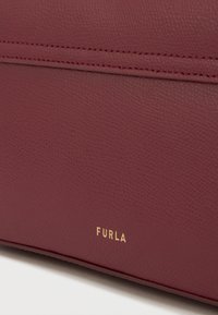 Μπούρντι δερμάτινη τσάντα με υφή, διαθέτει χρυσό λογότυπο "FURLA" και απλές λεπτομέρειες ραφής κατά μήκος των ραφών.