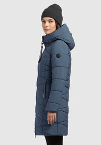 Modrý páperový kabát s kapucňou, s diagonálnymi prešívanými vzormi, zipsovými vreckami a nášivkou loga na rukáve.