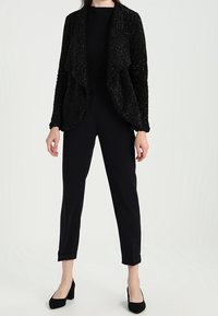 Černá pletená bunda s překládaným předním dílem, nošená přes sladěný top a zúžené kalhoty. Vyznačuje se hladkou texturou a přiléhavým střihem.