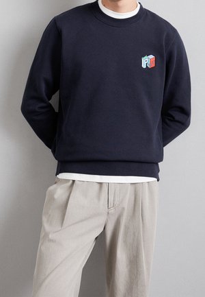 Felpa navy con scollo rotondo, caratterizzata da un piccolo logo geometrico multicolore sul petto. Indossata sopra una camicia bianca.