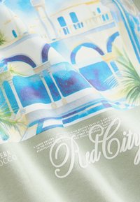 Pamučna majica s printom šarene arhitekture u plavoj i bijeloj boji s zelenim naglascima. Tekst glasi "Red City" u kurzivnom fontu.