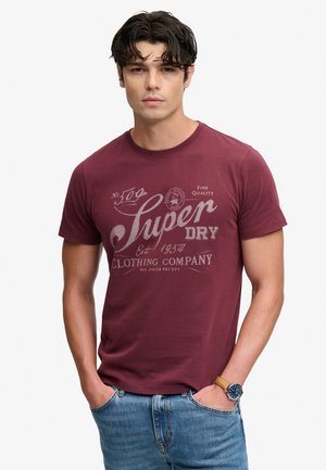 Ein junger Mann mit dunklen Haaren trägt ein weinrotes Superdry-T-Shirt und blaue Jeans, die Hände in den Taschen und eine Uhr am linken Handgelenk.