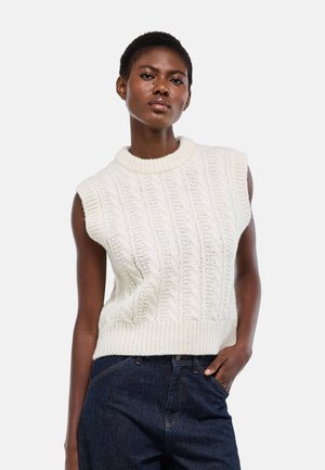 Femme portant un pull sans manches blanc à torsades et un jean bleu foncé, une main dans la poche, debout devant un fond uni.