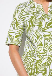 Camisa de patrón verde y blanco con mangas cortas, que presenta un diseño de cuello en V y una mezcla de formas de hojas y abstractas a lo largo de la tela.