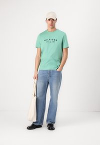 Grønn bomull t-skjorte med svart logo, lyseblå jeans, svarte sko og en beige caps. Holder en beige totebag. Enkelt, uformelt utseende.