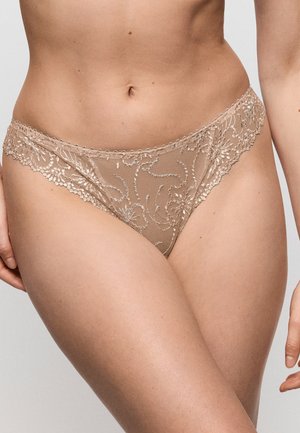 Vrouw die beige kanten bikini-ondergoed met bloemenborduursel draagt, getoond van taille tot bovenbenen tegen een effen achtergrond.