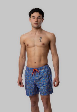Giovane uomo in piedi senza maglietta, indossa pantaloncini da bagno blu a fantasia con coulisse arancione, rivolto in avanti su uno sfondo grigio uniforme.