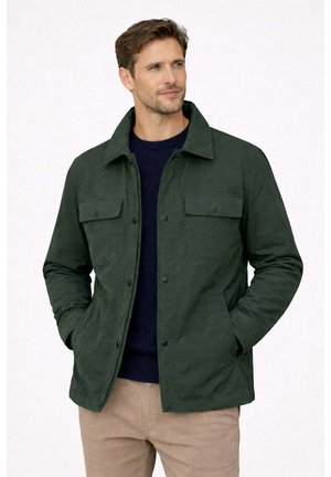 Uomo che indossa una giacca verde scuro con bottoni e tasche a toppa, maglione blu navy e pantaloni beige, in piedi con le mani nelle tasche, guarda di lato.