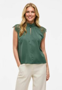 Zielona bezrękawnikowa bluzka z koronkowymi akcentami na ramionach, dekoltem w kształcie łezki i gładką teksturą, w zestawie z jasnymi spodniami.