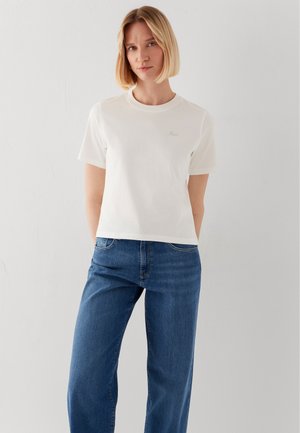 Frau mit blondem Bob-Haar, die ein schlichtes weißes kurzärmeliges T-Shirt und blaue Jeans trägt und vor einer weißen Wand steht.