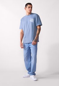 Lyseblå bomull T-skjorte med et lite hvitt grafisk motiv på venstre bryst, kombinert med avslappede lyseblå jeans og hvite joggesko.