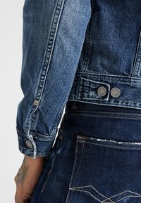 Jeansjacka med blåtvätt, med knappmanchetter, slitna detaljer och kontrasterande sömmar vid midjan och fickorna.