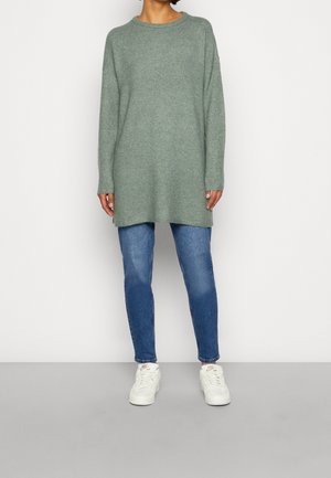 Person trägt einen langen grünen Strickpullover, blaue Jeans und weiße Nike-Sneaker und steht vor einem einfarbigen, hellen Hintergrund.