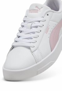 Białe skórzane sneakersy z różowym logo Pumy, teksturowana powierzchnia i płaskie białe sznurowadła. Posiadają zaokrąglony nos i gumową podeszwę z wzorami antypoślizgowymi.