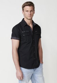 Camisa negra de manga corta con botones, con acentos de color gris en el cuello y dos bolsillos en el pecho, hecha de una tela ligera; ajuste casual y textura suave.