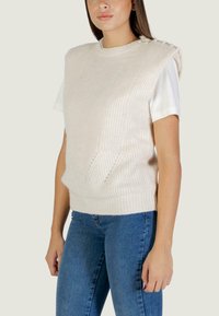 Gilet lavorato a maglia color crema con texture a coste, scollatura rotonda e bottoni laterali, indossato sopra una camicia bianca a maniche corte e jeans blu.