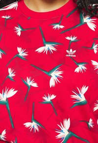 Maglietta rossa Adidas con motivo di fioritura tropicale uccello del paradiso bianco e verde su tutto il tessuto.