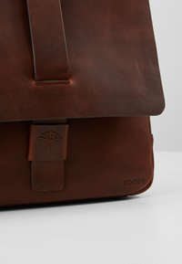Hnědá kožená taška přes rameno se strukturovaným designem, obsahuje klopu na zavírání, embosované detaily a decentní logo značky na boku.