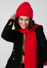 Bonnet en tricot rouge à côtes, écharpe rouge, manteau noir texturé avec des accents de boutons dorés, et pull beige clair. La personne semble joyeuse.