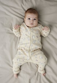 Bjelkasti zip-up kombinezon s uzorkom zečeva. Mekani pamuk, duži rukavi i elastične manžete. Pozadina je svijetlosmeđa tkanina.