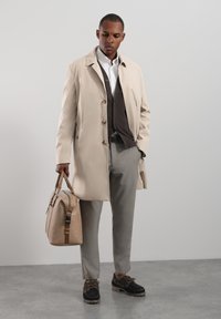 Abrigo beige tipo gabardina con botones marrones, sobre un chaleco oscuro y una camisa blanca, combinado con pantalones gris claro y zapatos oscuros, sosteniendo un bolso beige.