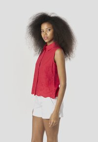 Rode mouwloze blouse met een knoopsluiting aan de voorkant, een geschulpte zoom en bloemenborduursels, gecombineerd met witte korte broek.