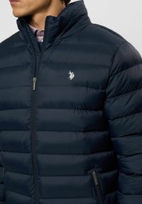 Mees, kes kannab mereväe sinist tikitud jopet, millel on ees tõmblukk ja rinnal valge Polo mängija logo, kandes selle all kraega särki.