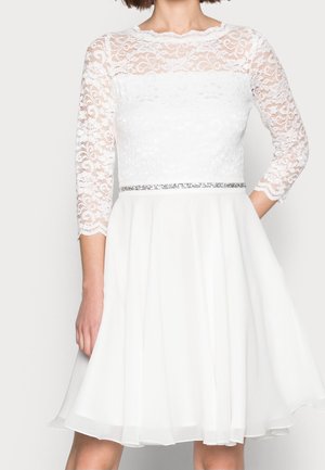 Femme portant une robe blanche mi-cuisse avec des manches longues en dentelle et un détail de ceinture argentée à la taille.