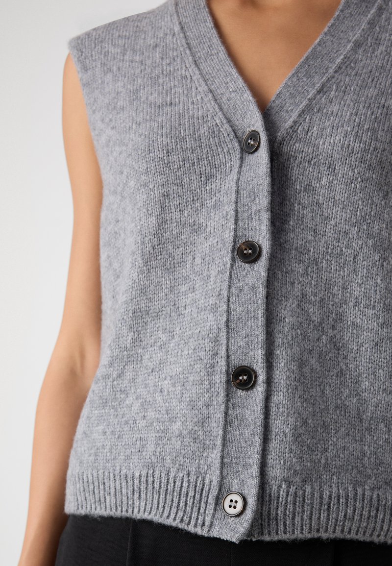 Grau gestrickter Pullover mit V-Ausschnitt, Knopfverschluss und geripptem Saum. Verfügt über dunkle Knöpfe und eine weiche Textur, ohne Ärmel gestaltet.