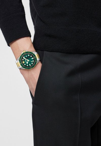 Személy fekete pulóvert és nadrágot visel, bal kezét a zsebében tartja, és két tónusú, zöld számlapos, arany részletekkel díszített fém karórát mutat.