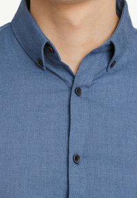 Camicia button-up in tessuto azzurro chiaro con una trama texture, caratterizzata da un colletto classico e bottoni neri lungo la pattina.