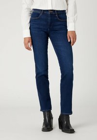 Donkerblauwe denim jeans met een slim fit, voorzien van een klassieke vijf-pocketontwerp, subtiele stiksels en een hoge taille. Draag het met zwarte haklaarzen.