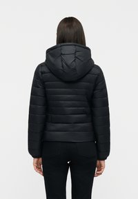 Γυναίκα με μαύρο καπιτονέ μπουφάν με κουκούλα, σε όρθια στάση με την πλάτη στραμμένη, μπροστά σε απλό ανοιχτό φόντο.