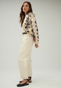 Pull beige avec motif floral noir, pantalon large crème, et chaussures plates noires avec des lanières. Texture lisse et coupe décontractée.