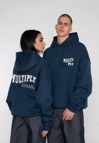 Navy blue hoodies feature front pockets, oversized fit, and embroidered "MULTIPLY" and "APPAREL" texts on the back. Soft material, casual design.