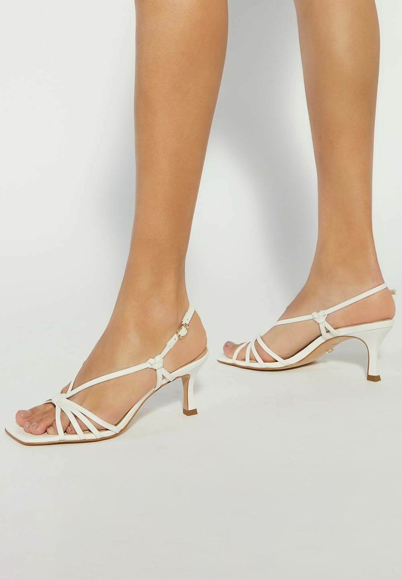 White strappy sandals with a narrow heel. Made of smooth material, featuring a unique knot detail and an adjustable ankle strap.