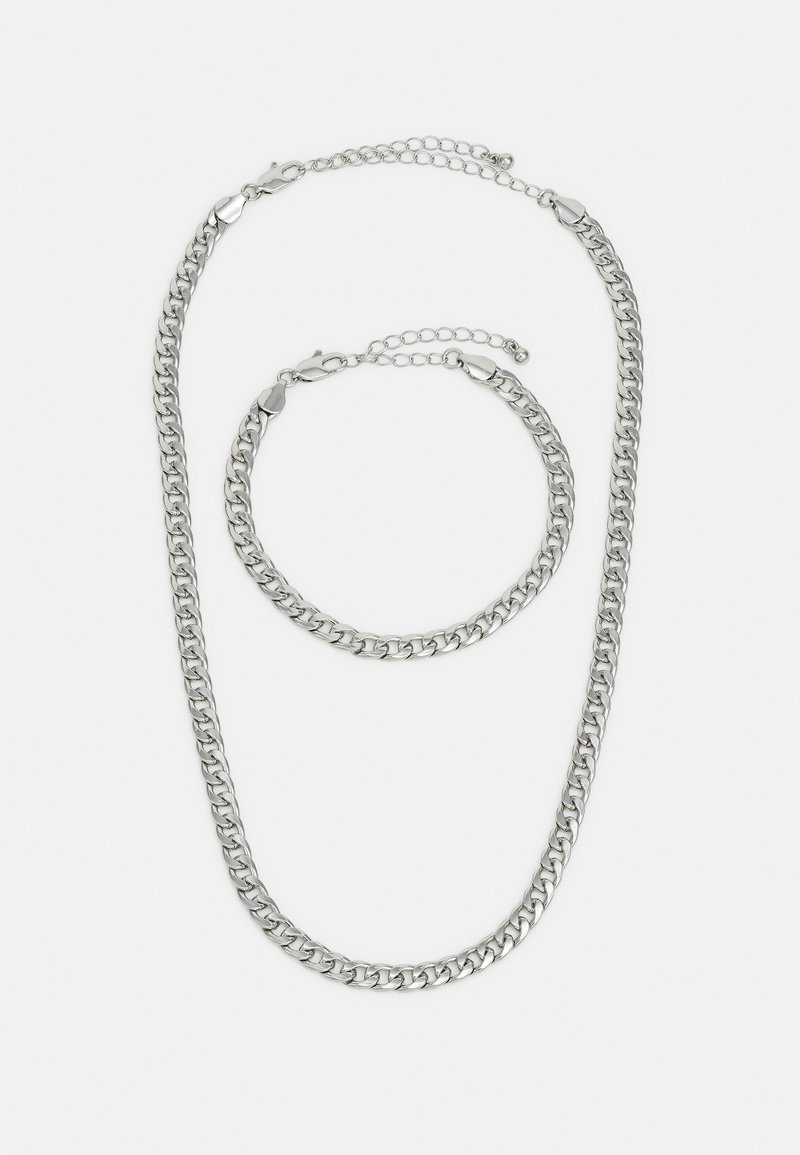 Conjunto de colar e pulseira de corrente de metal prateado, com elos grossos de textura lisa e fechos de mosquetão, dispostos em estilo sobreposto.