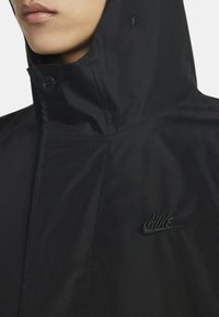 Giacca Nike nera con colletto alto e cappuccio, dotata di un logo testurizzato ricamato sul lato sinistro del petto e una finitura in tessuto liscio.