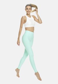 Fehér sportmelltartó, magas derekú menta színű leggings sima, rugalmas anyagból. Modell mezítláb sétál, haja lófarokba fogva, mosolyog.