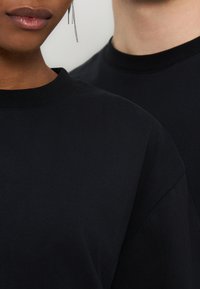 Δύο άτομα φοράνε μαύρα μπλουζάκια με στρογγυλή λαιμόκοψη, το ένα με μεσαίου τόνου καφέ επιδερμίδα και ένα εμφανές κρεμαστό σκουλαρίκι αλυσίδας.