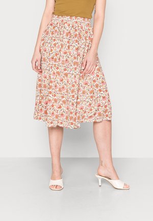 Vrouw draagt een midi bloemenrok met oranje en roze bloemen, gecombineerd met witte muiltjes met hak en een mosterdkleurige top.
