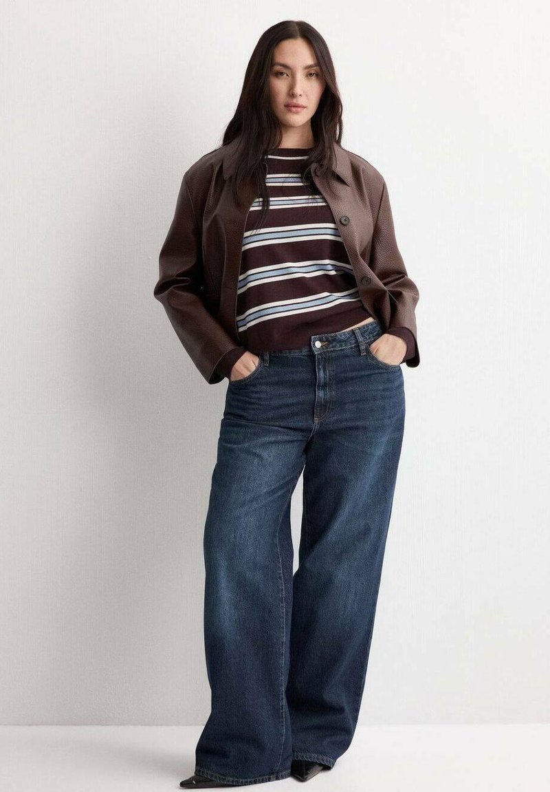 Femme debout, les mains dans les poches, portant une veste en cuir marron, une chemise rayée, un jean large bleu et des chaussures noires à bout pointu, devant un mur uni.