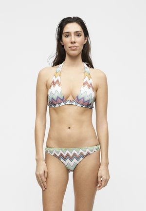 Kvinde med langt mørkt hår iført en flerfarvet bikini med zigzag-mønster og halterhals, stående mod en ensfarvet lys baggrund.