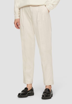 Persoon die off-white geplooide broek en zwarte glanzende loafers draagt, staand met één hand in de zak tegen een lichte achtergrond.