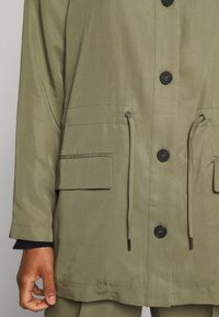 Veste verte à manches longues avec boutons à l'avant, cordons à la taille, poches à rabat, portée sur une manche noire avec bord-côte, main reposant sur le côté.
