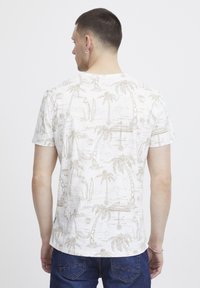 Camiseta de algodón blanca con estampado tropical en beige claro, que presenta palmeras y tablas de surf. Mangas cortas y cuello redondo.