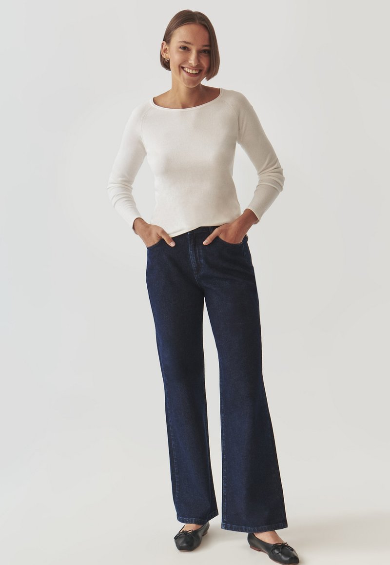 Weißer Pullover mit langen Ärmeln und weitem Ausschnitt, kombiniert mit dunkelblauen Schlagjeans und schwarzen Schuhen. Das Modell steht mit den Händen in den Taschen.
