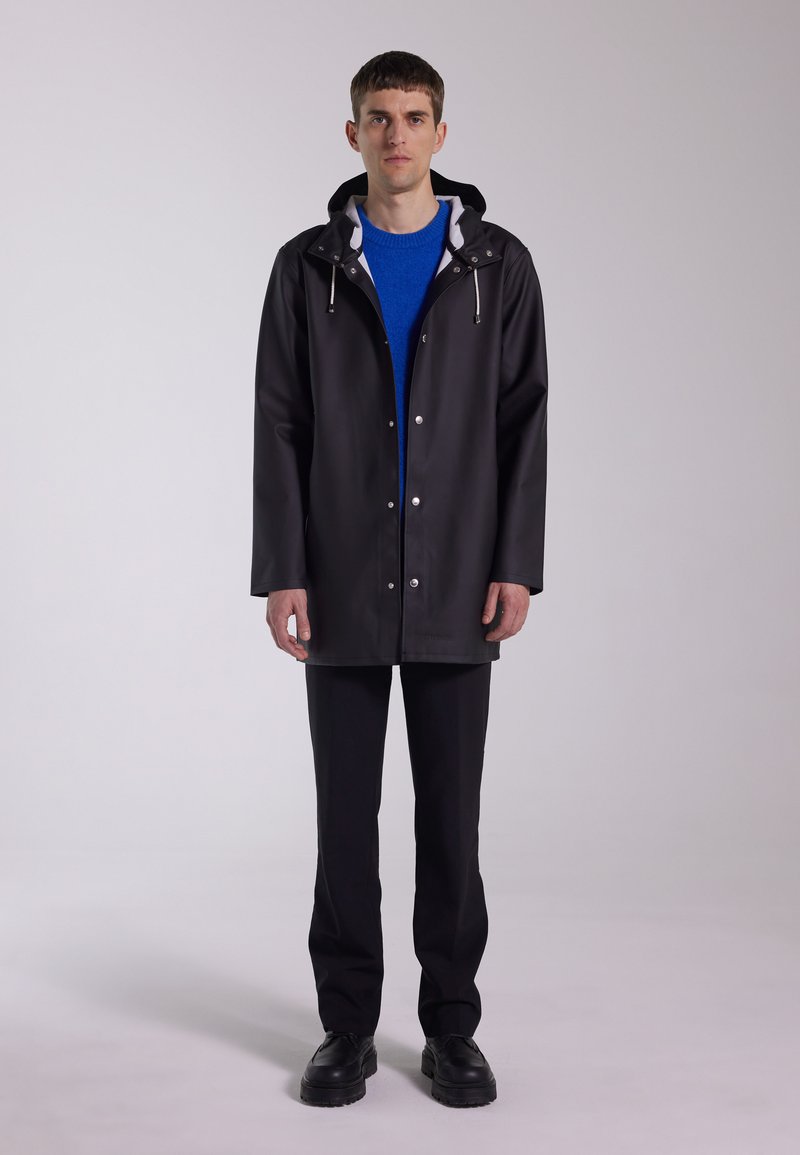 Svart vattentät regnrock med huva, tryckknappar och dragsko. Buren över en blå tröja och kombinerad med svarta byxor och skor.