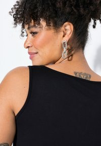 Czarny bezrękawnik o gładkiej teksturze. Posiada okrągły dekolt i srebrne kolczyki w kształcie łańcuszka. Widoczny tatuaż na górnej części pleców.